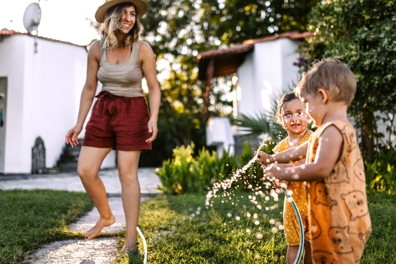 Frau mit zwei Kleinkindern im Sommer im Garten, die mit einem Wasserschlauch spielen