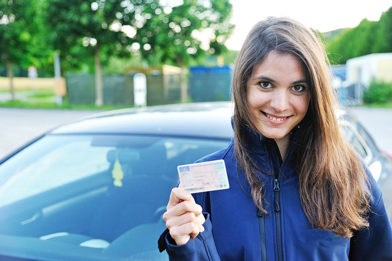 Junge Fahranfängerin hält lachend ihren Führerschein in der Hand. 